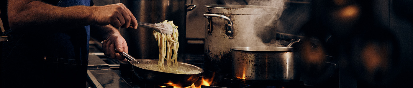 Commercial kitchen with steaming pots and creamy spaghetti being pulled out of a pan